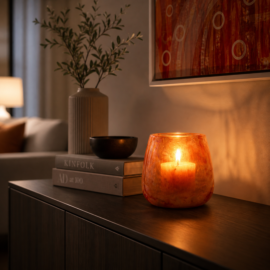 Modernes Windlicht aus orangefarbenem Glas mit brennender Kerze auf einer Kommode in stilvoll eingerichtetem Wohnzimmer – warme Lichtstimmung und elegante Dekoration