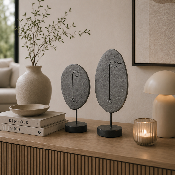 Zwei abstrakte Gesichtsfiguren in Grau-Schwarz als modernes Deko-Set auf einer Holzkommode in einem stilvoll eingerichteten Wohnzimmer mit minimalistischen Wohnaccessoires und warmem Licht.
