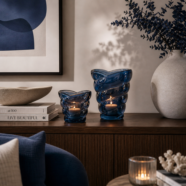 Zwei blaue Windlichter aus Glas in unterschiedlicher Größe dekorativ auf einer Holzkommode in einem modern eingerichteten Wohnzimmer mit stilvollen blauen Akzenten