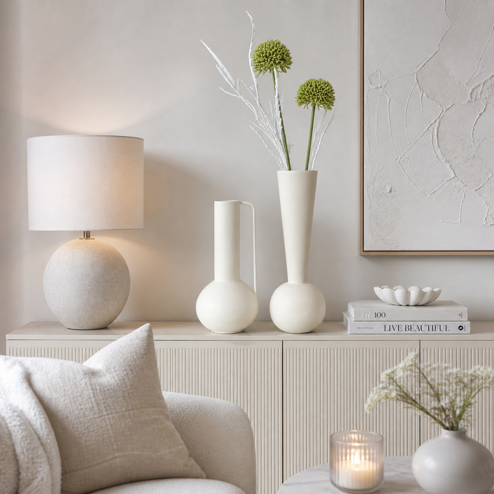 Zwei moderne Metallvasen in creme matt, eine schlank und eine bauchig, dekoriert auf einer Kommode in einem hellen, stilvoll eingerichteten Wohnzimmer mit weißen und cremefarbenen Akzenten