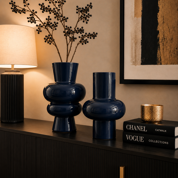 Zwei moderne dunkelblaue Metallvasen in unterschiedlichen Größen auf einem Sideboard in einem stilvollen Wohnzimmer mit warmer Beleuchtung und minimalistischer Einrichtung
