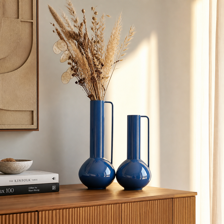 Zwei elegante blaue Metallvasen auf einem Holz Sideboard, dekoriert mit Trockenblumen in einem modernen minimalistischen Wohnzimmer mit warmem Licht