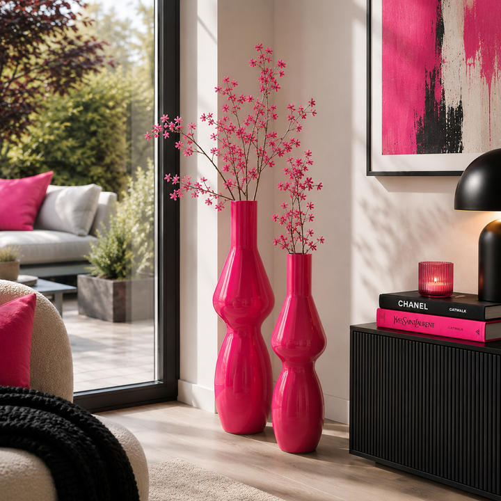 Zwei glänzende pinke Bodenvasen mit dekorativen Zweigen in einem stilvoll eingerichteten Wohnzimmer vor einem großen Terrassenfenster mit modernen schwarzen und pinken Akzenten.